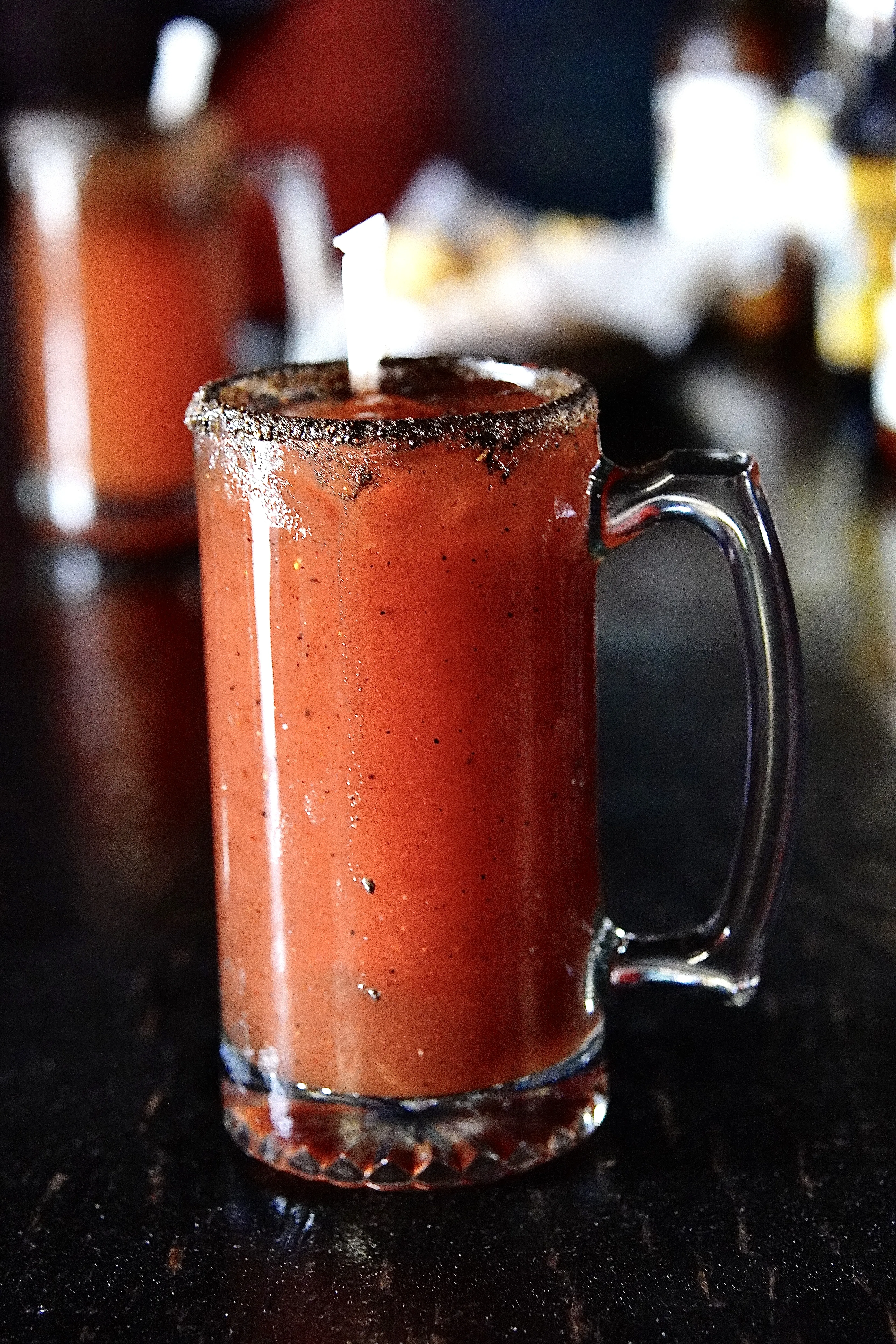 Red Chili covered Michelada. The brim is chili covered with a blurry background of another michelada and beers.