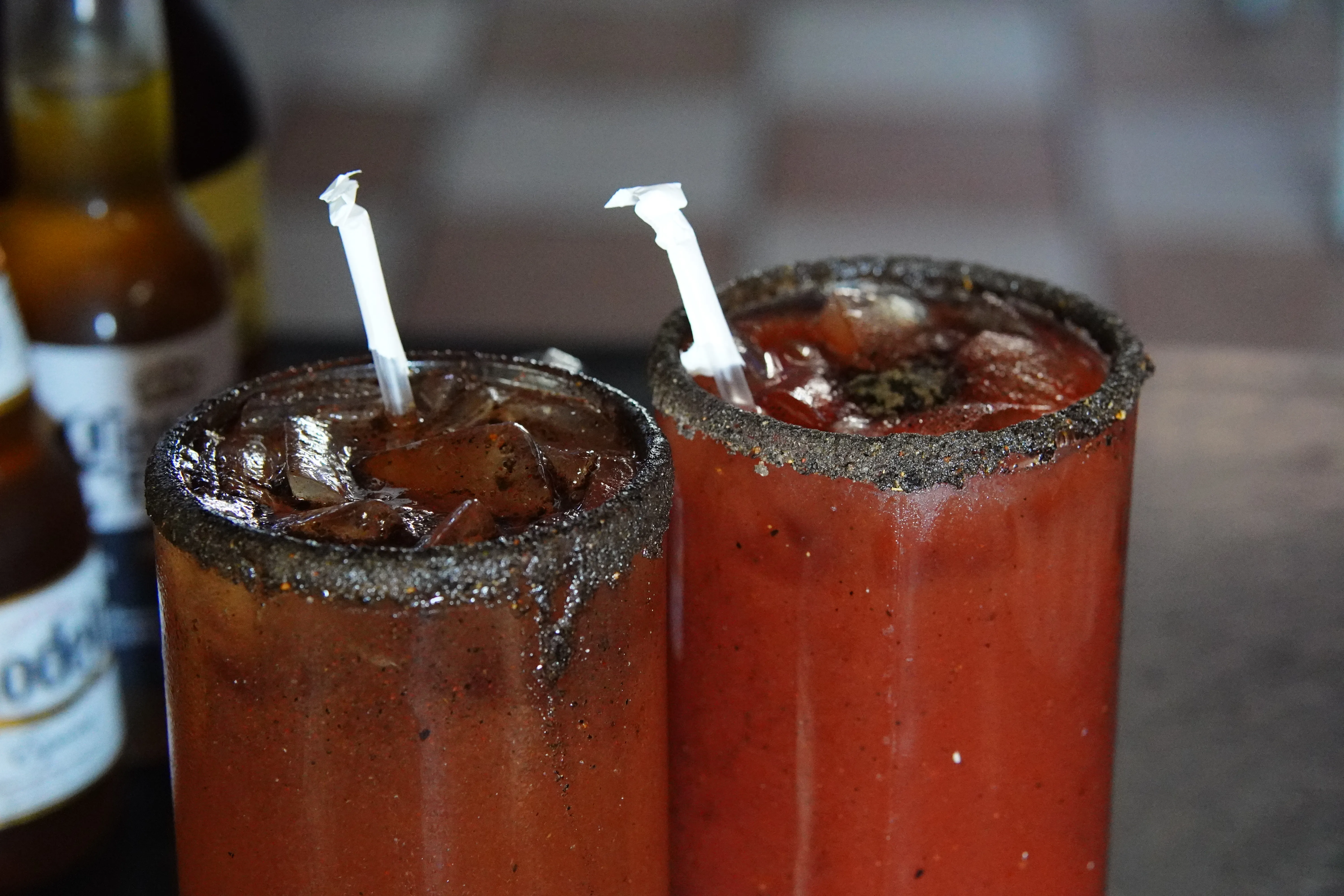 The tops of 2 ice cold micheladas. the cup is overflowing and seating because of how cold  it is with ice poping out of the top. 3 beers in the background.